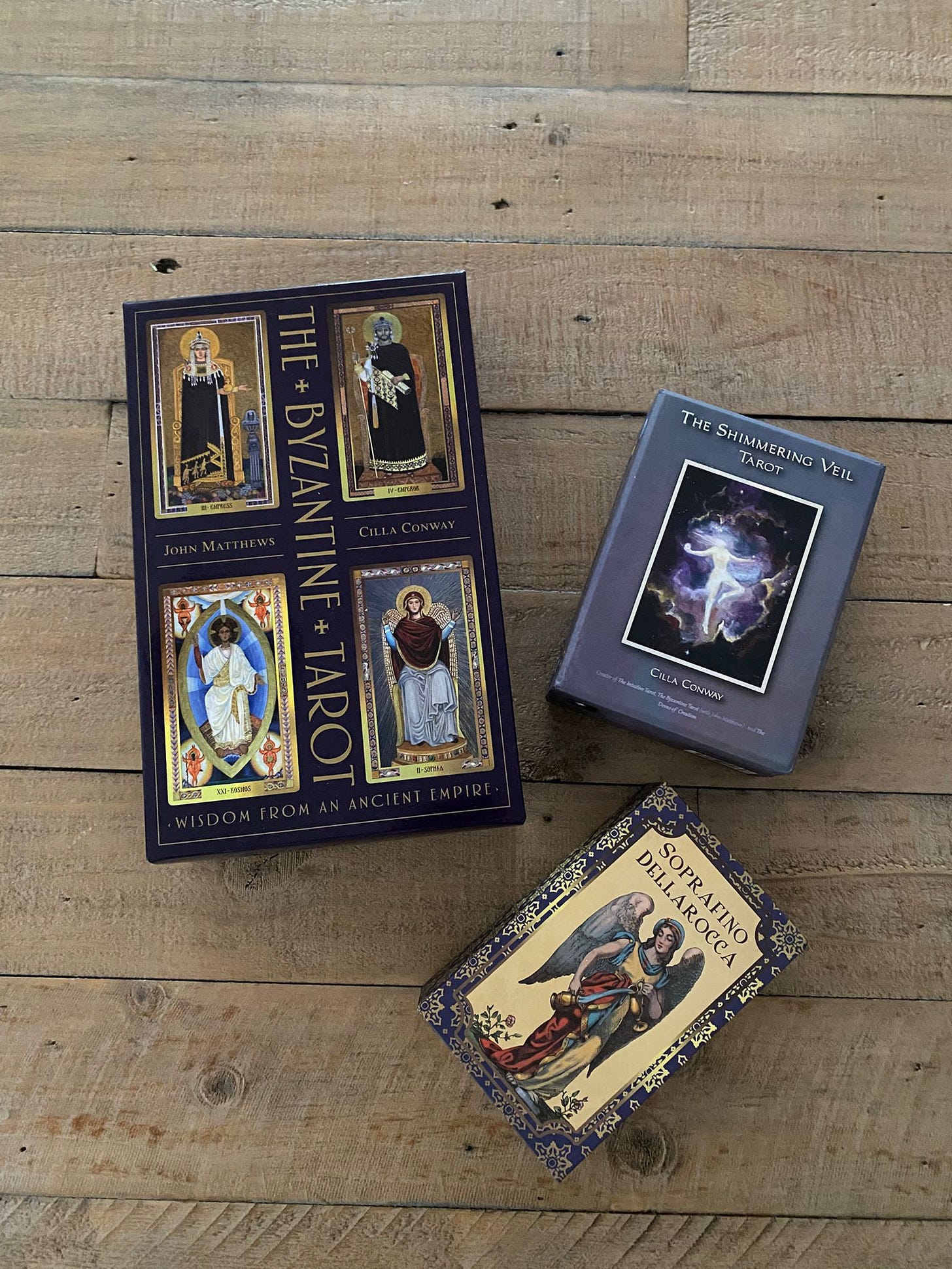 Three tarot and oracle decks arranged on a wooden floor for a Tarot DMs interview with Emma van Straaten, including The Byzantine Tarot, The Shimmering Veil Tarot, and Soprafino della Rocca, showing the eclectic visual references behind the conversation.