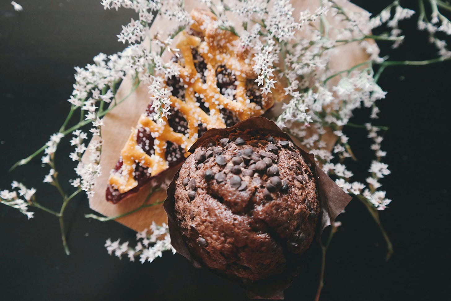 lattice-top pie on dark background next to a chocolate muffin in case, small white flowers lay across both.