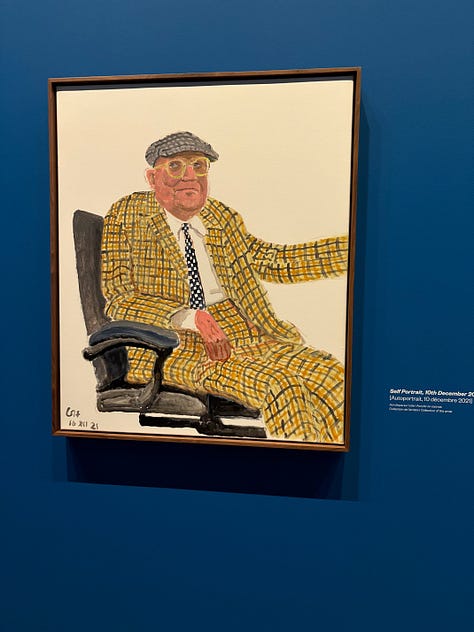 Multiple bright-colored paintings including a self-portrait of Hockney in a yellow plaid suit and big yellow glasses, the crowd viewing paintings, a mother and child in front of a bright painting of a massive tree, an annunciation painting redone in neon colors, a green lawn with sprinkler spray, a photograph of hockney working at his drawing table with his little terrier dog under the table and two paintings with raindrops hitting puddles among trees