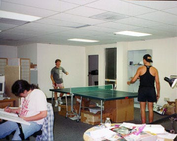 Two men playing ping pong, one working at a drawing board