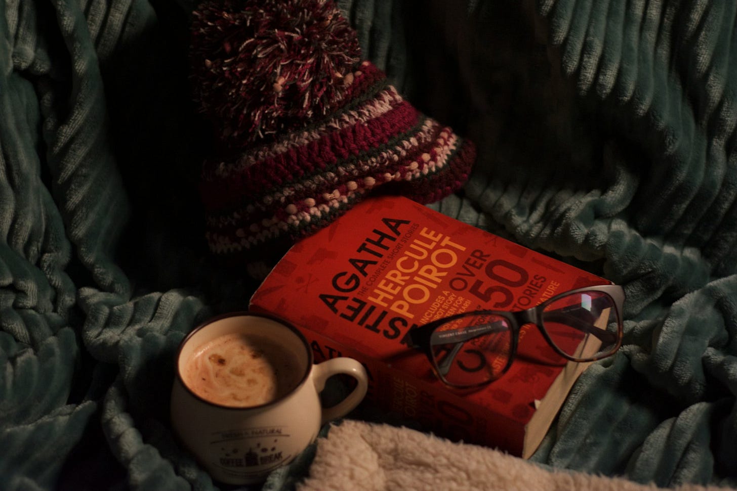 A large Agatha Christie collection with a red cover rests on a teal green corduroy blanket, along with a mug of hot cocoa and a deep red, white, and olive green knit hat with a pompom top.