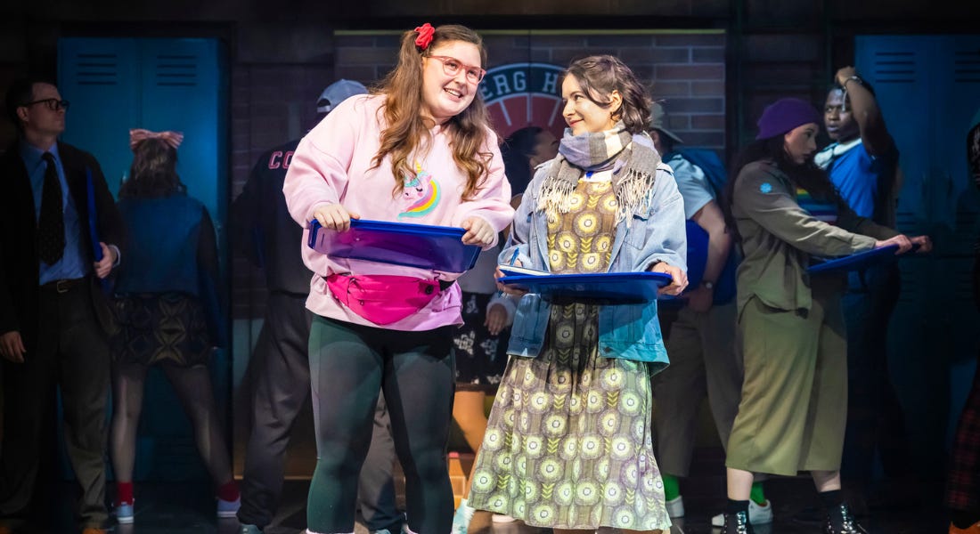 Two people on stage - one wearing a pink jumper and leggings, and one a green dress and blue scarf Two people on stage - one wearing a pink jumper and leggings, and one a green dress and blue scarf