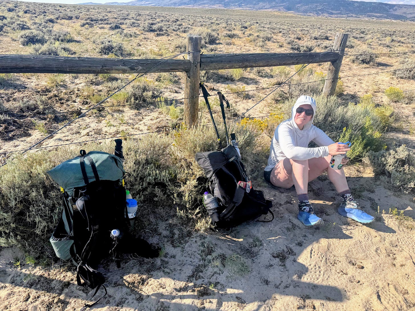 Author sits half in shade on sand and sagebrush beside a fence in the middle of the Great Divide Basin in southern Wyoming. There are many, many trail runner footprints visible in the sand. 