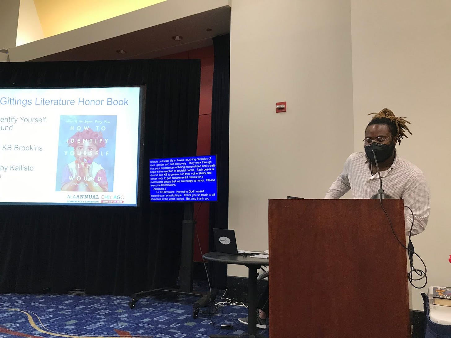 KB speaking at a podium as their book, "HOW TO IDENTIFY YOUSELF WITH A WOUND" and text detailing their award shows in the background.