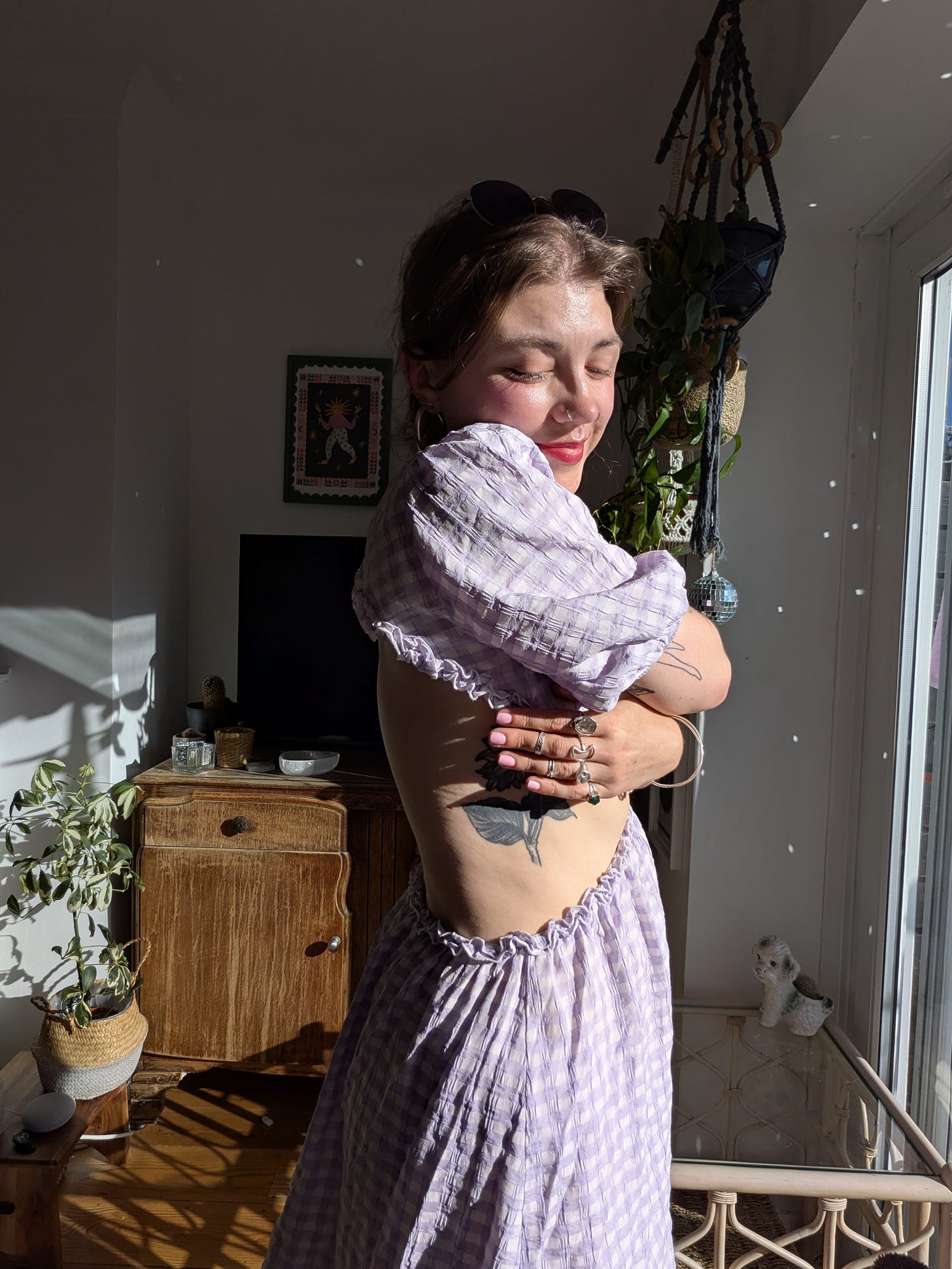 Grace hugging herself in the sunlit living room (disco ball reflection speckles visible on the back wall), and she's wearing a ruched (sp?) gingham dress (lilac and white checks) with cut-out sections at the back so it reveals the sunflower tattoo on her ribs