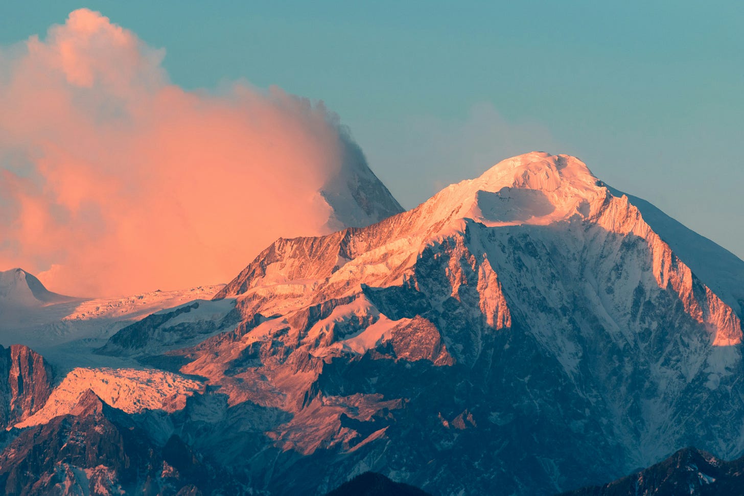 Snow-covered mountain peaks bathed in warm pink and orange at sunset, with clouds billowing behind the summit against a teal blue sky.