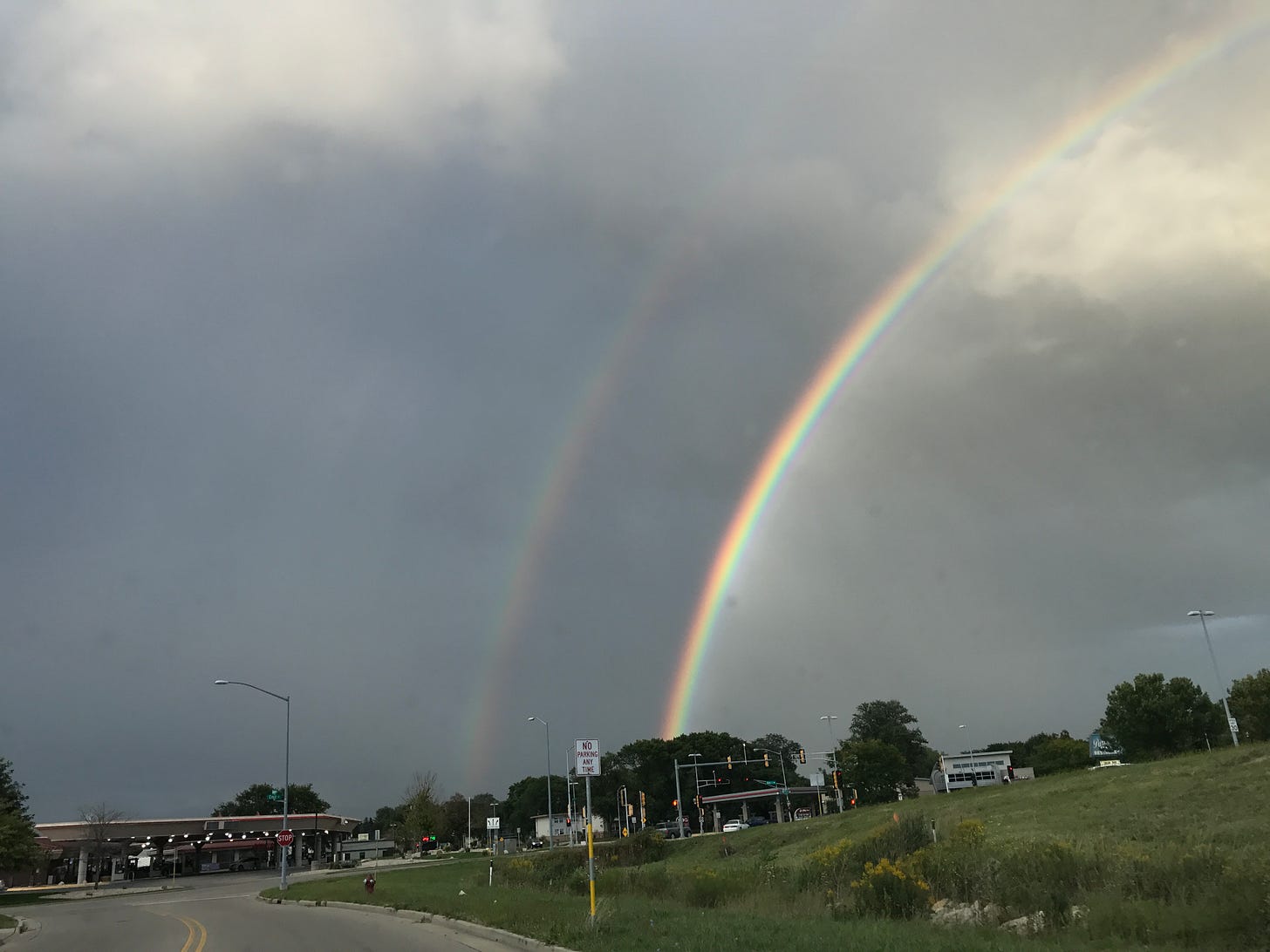 A double rainbow arcs across a gray sky, partially lit by evening sun. The photo is taken from inside a car approaching an intersection, with green grass and roadside signs in the foreground. Captured on September 5, 2017, while driving to Michael Piechowski’s home for the first evening together.