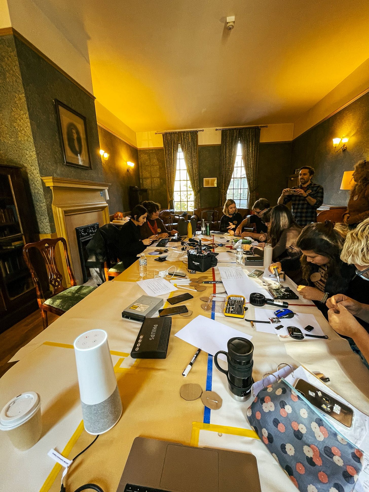 room full of people sat around a large oval table. Table is covered in paper and broken electricals as the people play with the materials room full of people sat around a large oval table. Table is covered in paper and broken electricals as the people play with the materials