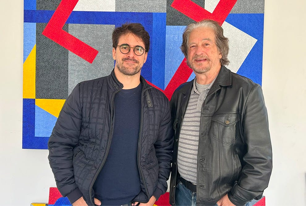 Duas pessoas em pé lado a lado diante de um painel de arte abstrata composto por formas geométricas em azul, vermelho, amarelo, cinza e branco; a pessoa à esquerda veste jaqueta acolchoada preta sobre camiseta azul-marinho e óculos de armação redonda, enquanto a pessoa à direita veste jaqueta de couro cinza sobre camisa listrada em tons neutros, ambas com as mãos relaxadas e olhando para a câmera.