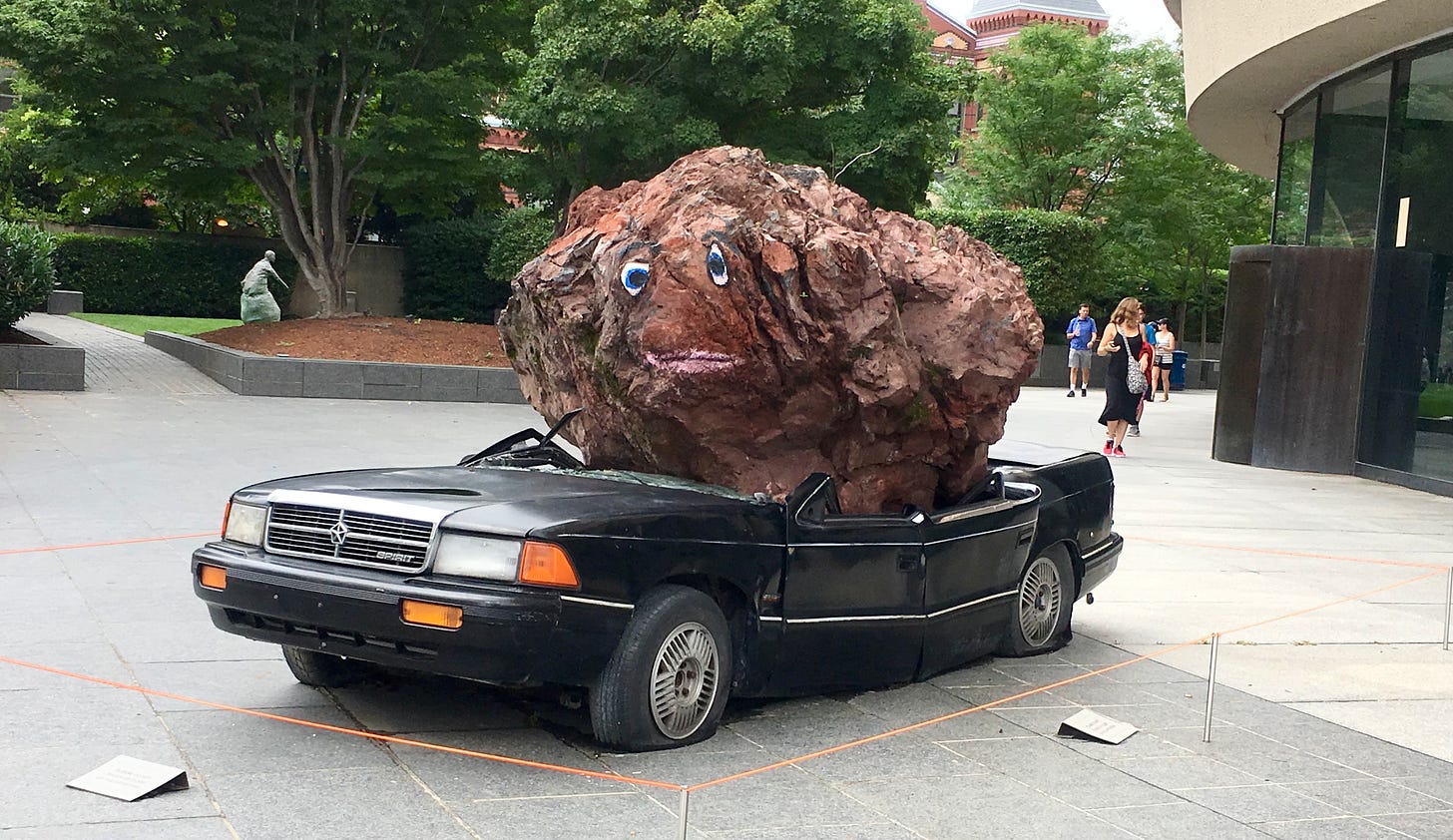 a full-size 1970s car with a massive rock with googly eyes, dropped on top of it