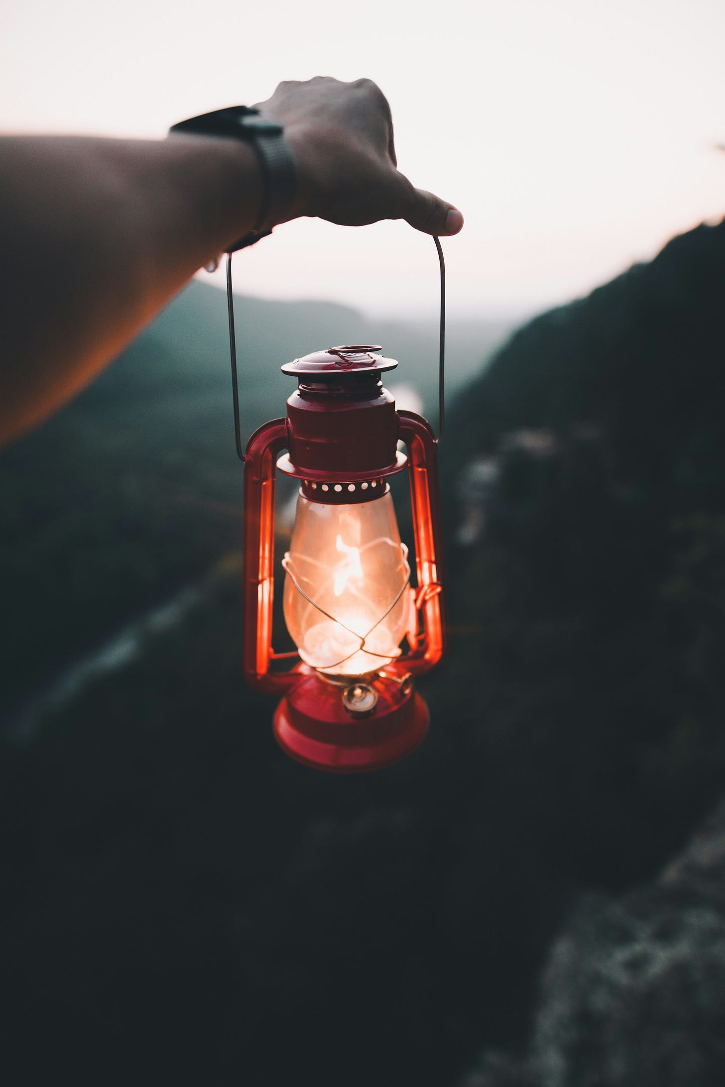 A person’s outstretched arm holding a red lantern glowing with warm light, suspended over a mountainous landscape at dusk. The background is softly blurred, suggesting depth and distance, evoking a sense of journey, guidance, and illumination. A person’s outstretched arm holding a red lantern glowing with warm light, suspended over a mountainous landscape at dusk. The background is softly blurred, suggesting depth and distance, evoking a sense of journey, guidance, and illumination.