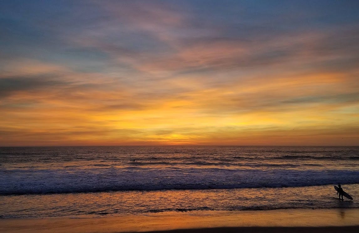 Picture taken just after sunset with a gold, pink, and blue sky reflected in the waves of the Pacific Ocean. A surfer stands in the shallows in the lower righthad corner, carrying their surfboard as they face the recently set sun