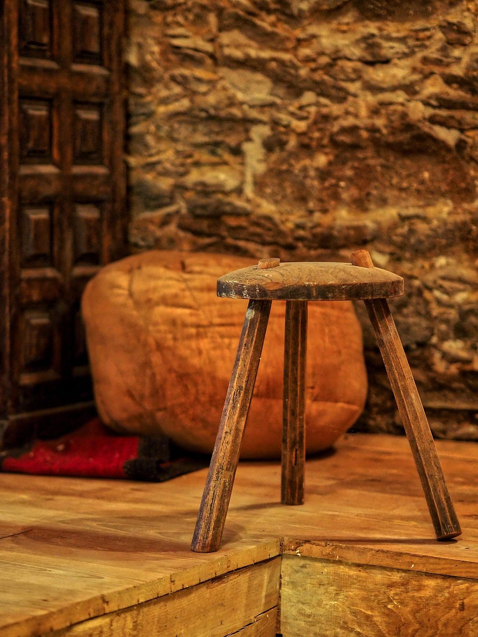 A picture of a rustic wooden three-legged stool sitting on a rough wooden floor, kind of uncomfortably near what appears to be an opening in said floor. Behind the stool is a burnt-orange bean back chair, an antique-looking paneled wooden door, and a rough stone wall. A picture of a rustic wooden three-legged stool sitting on a rough wooden floor, kind of uncomfortably near what appears to be an opening in said floor. Behind the stool is a burnt-orange bean back chair, an antique-looking paneled wooden door, and a rough stone wall.