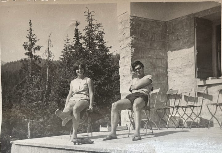 A black-and-white photo of a young couple in the late 1960s standing outdoors on a scenic hillside. The man, wearing sunglasses and a striped t-shirt with shorts, has his arm around the woman and is pointing toward the distance. The woman, wearing a sleeveless buttoned shirt and shorts, is smiling as she looks in the same direction. Tall evergreen trees and mountain peaks are visible in the background under a hazy sky.