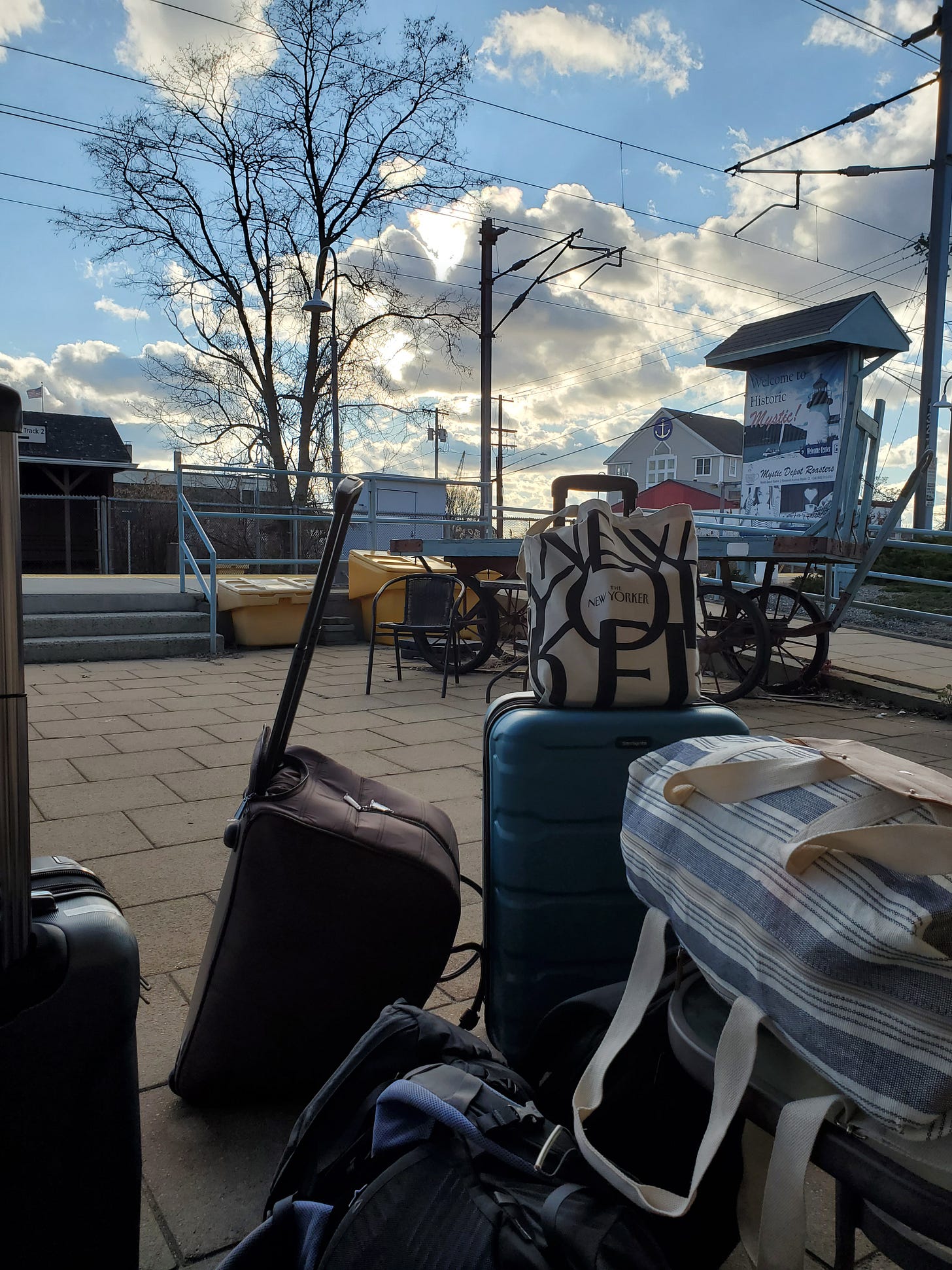 A mountain of luggage sits on an outdoor train platform with bare trees and bright blue sky, fluffy clouds behind. 