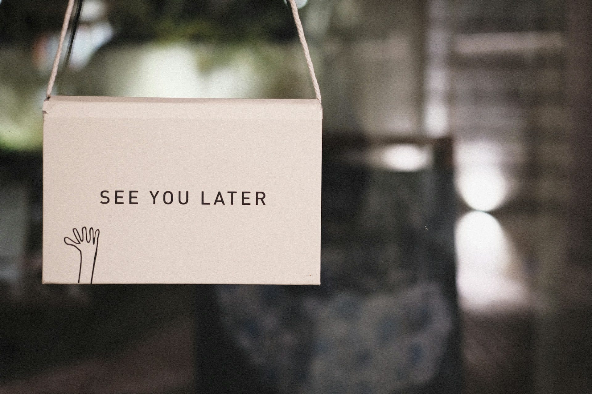 A white, rectangular hanging sign displaying the text 'SEE YOU LATER' in black capital letters, featuring a simple line drawing of a waving hand in the bottom left corner, set against a blurred, out-of-focus background A white, rectangular hanging sign displaying the text 'SEE YOU LATER' in black capital letters, featuring a simple line drawing of a waving hand in the bottom left corner, set against a blurred, out-of-focus background