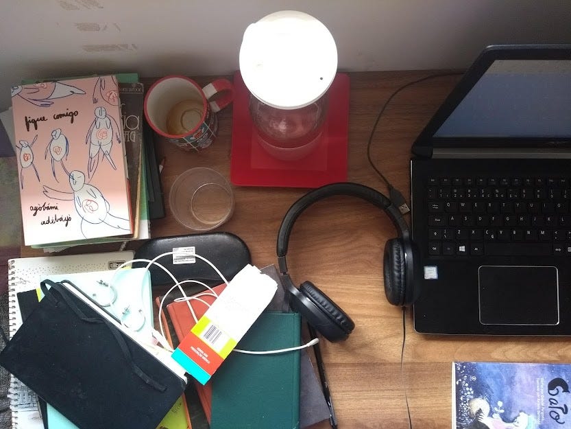 Imagem de uma mesa de trabalho vista de cima. A mesa é de madeira e está cheia de livros, cadernos, canecas sujas de café, copos, fones de ouvido e o computador está no canto.