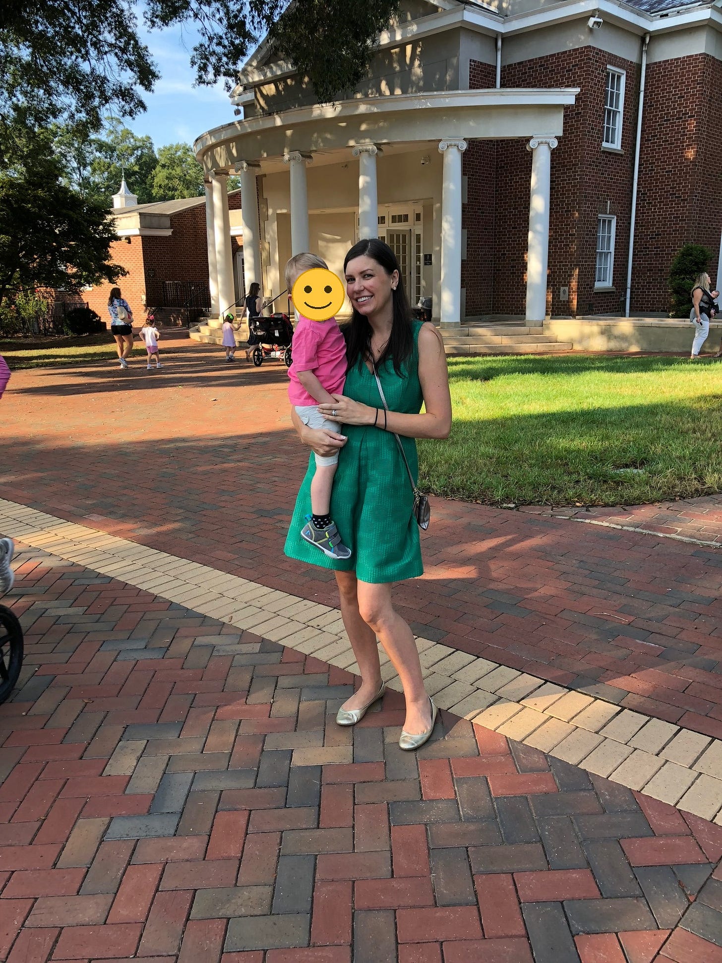 A woman in a green dress holds her young child on her hip at preschool drop-off on a brick walkway outside a columned building; a smiley emoji covers the child's face for privacy. A woman in a green dress holds her young child on her hip at preschool drop-off on a brick walkway outside a columned building; a smiley emoji covers the child's face for privacy.