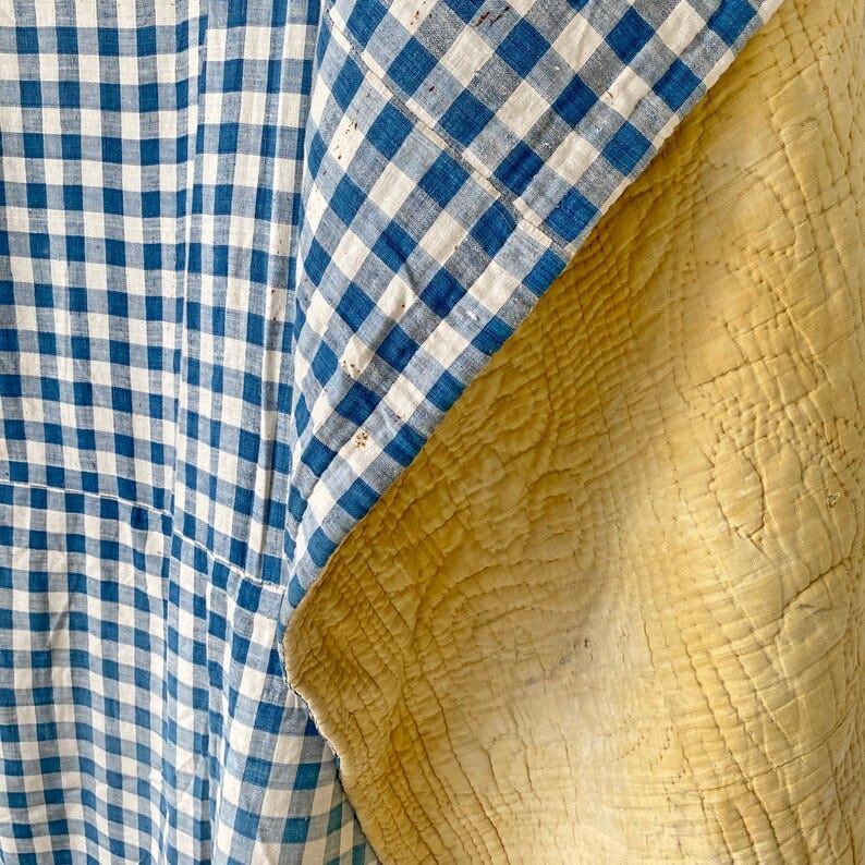 May include: A blue and white gingham fabric with a yellow fabric underneath. The gingham fabric is folded over the yellow fabric and the edges are visible. The yellow fabric has a quilted pattern.