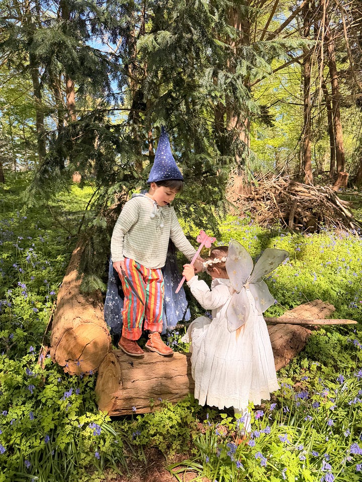Young girl picking flowers in a bluebell forest, and a boy helping her climb a log, both in costume