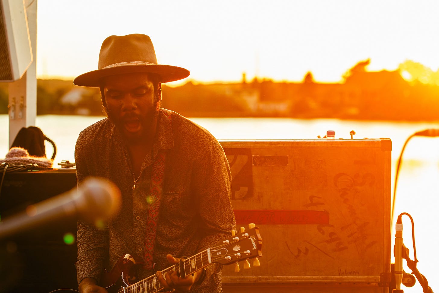 Gary Clark Jr. sings while playing his electric guitar on an outdoor stage. He is wearing a hat and is backlit by the bright, golden light of the setting sun over the water behind him.