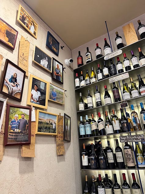 Photos inside In Vino wine bar in Yerevan, including a hand holding a bottle of Trinity Canyon Areni Noir, staff pouring wine behind shelves stocked with Armenian and international bottles, friends toasting with glasses of white wine, a long tasting table set with glassware beneath a sign reading 'The Renaissance of Armenian Wine Making,' and a wall of photos honoring winemakers