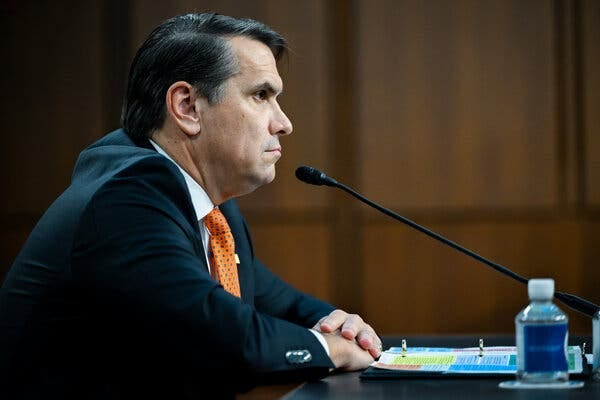 Blanche, in an orange tie and dark suit, sits behind a microphone.