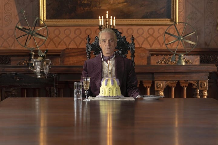 Jeremy Irons as Adrian Veidt sits at a long dining table in a lavish, old-world mansion, staring blankly ahead with a frosted purple-and-yellow cake lit by candles placed in front of him.