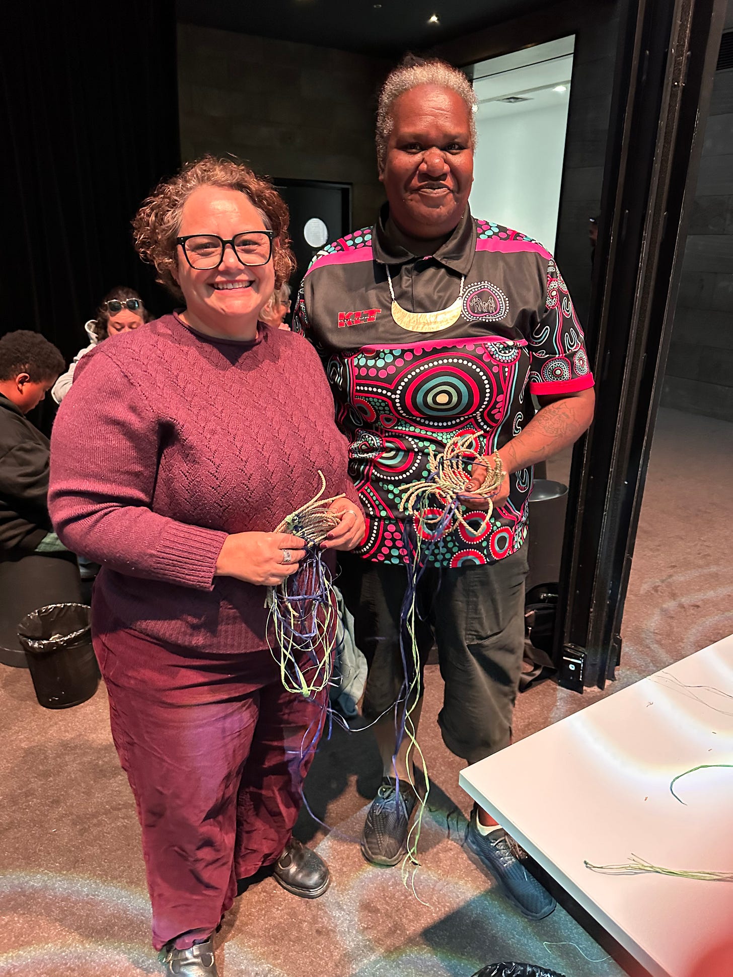 Two women stand side by side holding partially woven strands made from natural fibres. On the left, Lana Woolf wears a textured pink long-sleeve top and holds several thin, light-coloured strands in both hands. On the right, Paula Savage wears a black shirt with a bold, colourful circular pattern in red, green, and white, and holds a more complex bundle of woven and unwoven fibres, showing different stages of the weaving process. The image is partially obscured by a screen overlay at the top, suggesting it was taken during or after a workshop setting.