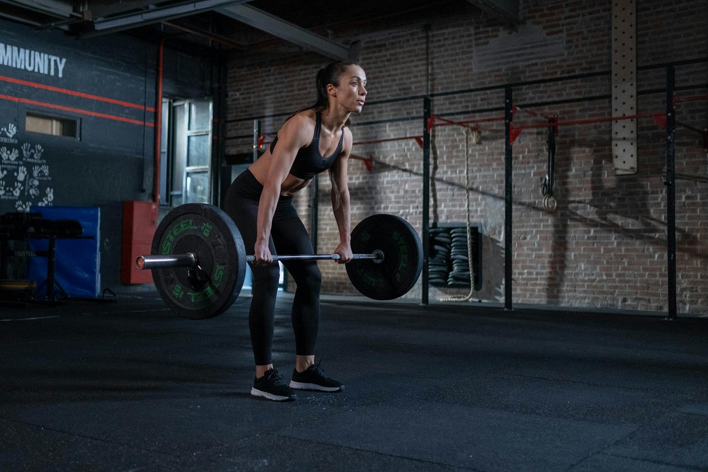 Woman in athletic wear deadlifting in a gym with brick walls. Focused expression, dim lighting, "COMMUNITY" text visible on left wall.