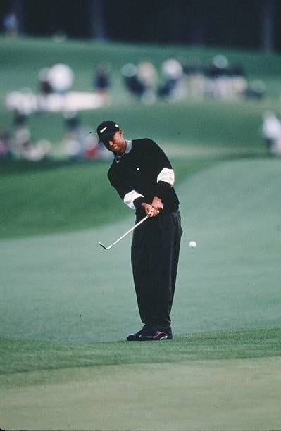 This may contain: a man in black shirt and pants playing golf on green with people watching from the sidelines This may contain: a man in black shirt and pants playing golf on green with people watching from the sidelines