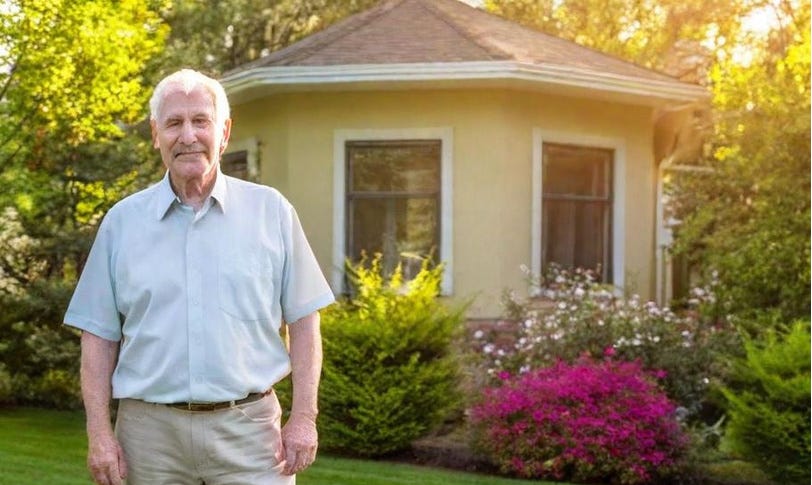 White-haired white man in light blue shirt and tan slacks stands in front of windowed six-sided porch with many green shrubs, one of which has bright-red flowers. White-haired white man in light blue shirt and tan slacks stands in front of windowed six-sided porch with many green shrubs, one of which has bright-red flowers.