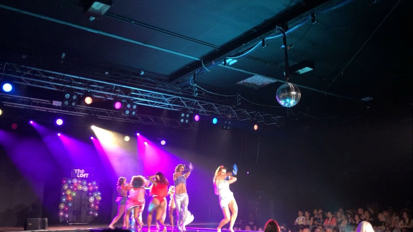 Photo of the house of suarez disco ball at Contact Theatre. Under bright pink lights, a collective house of performers vogue down the runway in a choreographed number. They wear sparkling 70s inspired outfits, and the crowd looks on cheering.