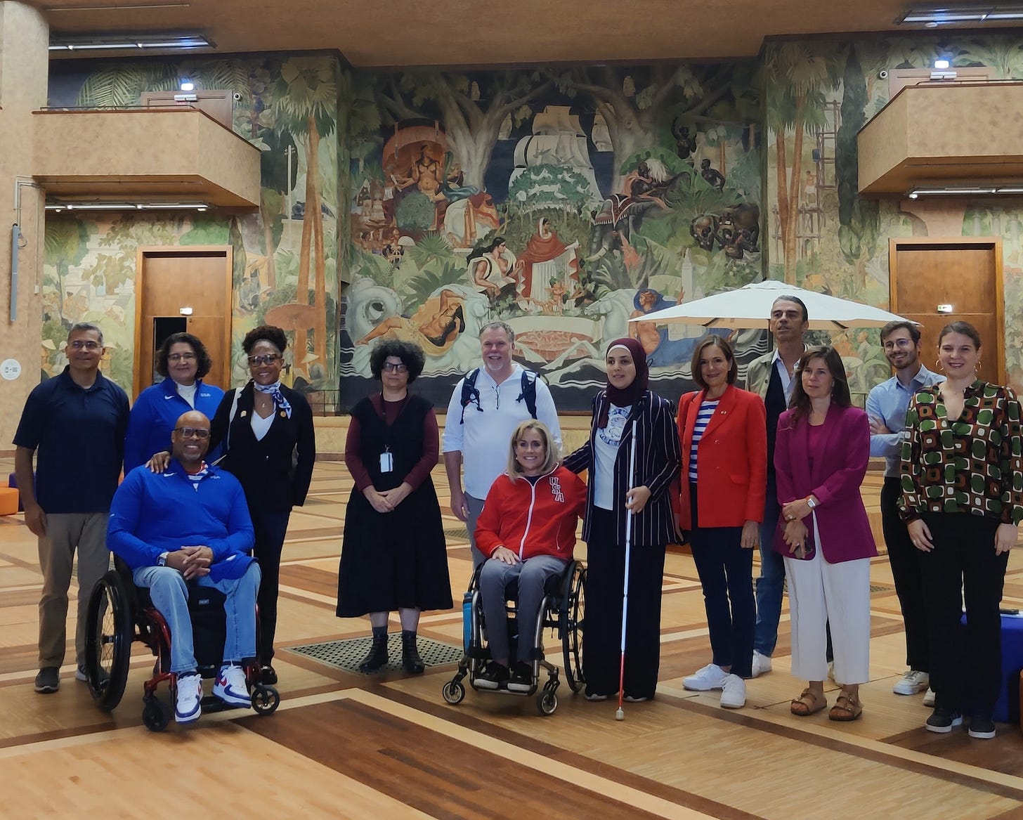 Sara stands at the center of a large group of people. She wears her official white Team USA t-shirt and a navy blazer with red vertical strips overtop. Her hand rests on the back of a woman in a wheelchair to her left, who wears a bright red Team USA jacket. 