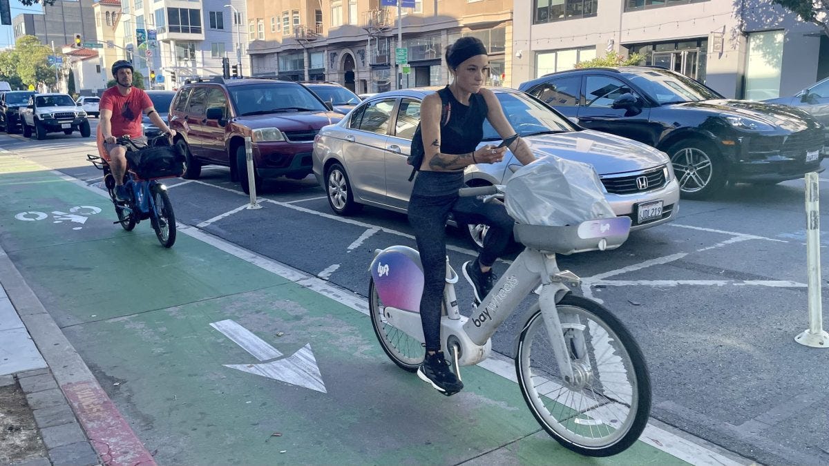 A woman rides a shared bike in a designated bike lane while looking at her phone. A cyclist follows behind her. Cars are parked and moving along the adjacent street. A woman rides a shared bike in a designated bike lane while looking at her phone. A cyclist follows behind her. Cars are parked and moving along the adjacent street.