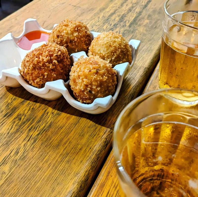 Photo of a wooden table, with two small cups of clear glass full of beer to the right, and to the left four balls of fried dough accompanied by a red sauce.