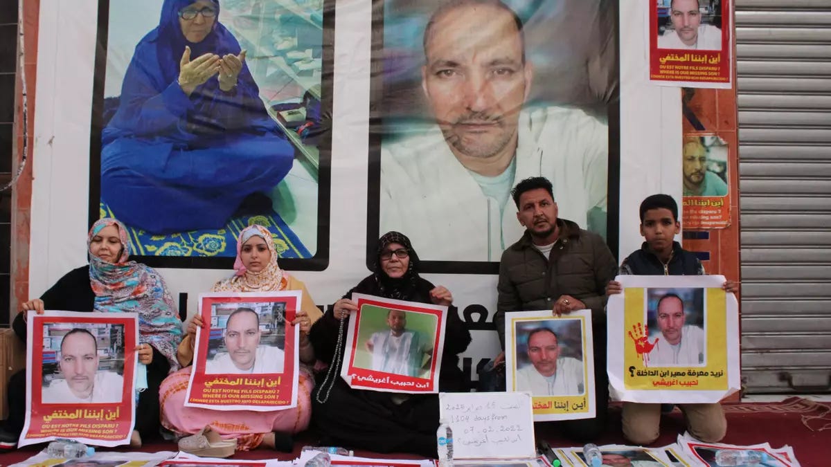  Tre donne e due uomini sono seduti o inginocchiati davanti a una fila di poster su un muro. I poster mostrano un uomo calvo con la barba corta. Una donna anziana in blu, la madre dell’uomo, con un hijab blu è visibile in un poster grande a sinistra, con le mani giunte in preghiera.