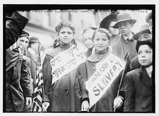 black and white image of two girls in early 1900s dress. There are boys in the background with hats. Girls are wearing sashes. One has writing in Hebrew and the other has "Abolish Ch... Slavery" 