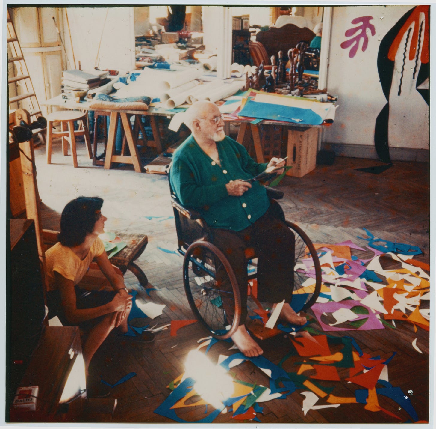 Matisse sitting in a wheelchair, surrounded by solorful cut-out scraps on the floor.