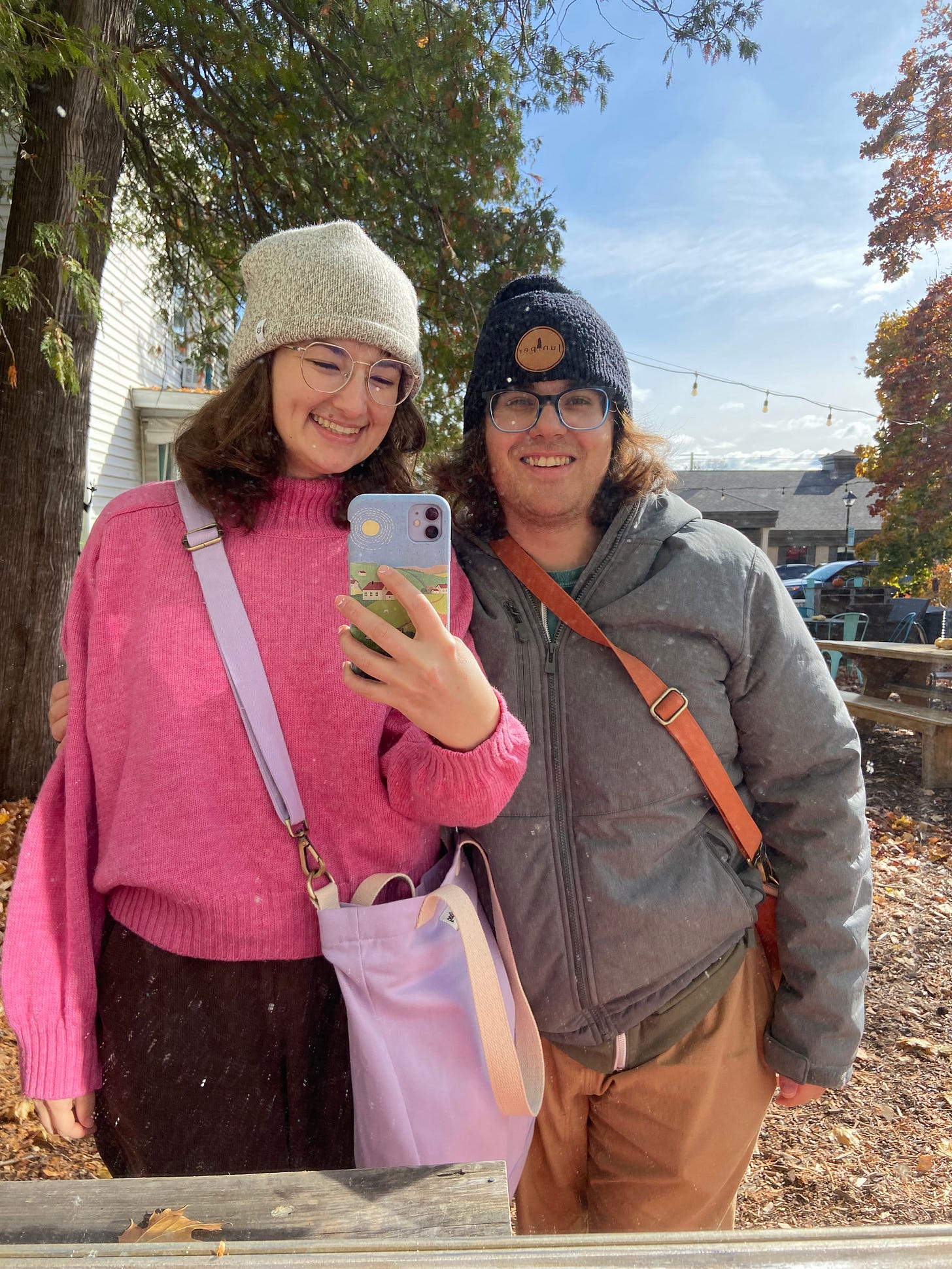 Ryan and Jenn smile and take a selfie in a mirror outside. It is fall in Sturgeon Bay, Wisconsin and there are trees with colorful leaves behind them. Jenn is wearing a white beanie, wire-rimmed glasses, a bubblegum pink turtleneck sweater, and brown pants. Ryan is wearing a black beanie, blue plastic-rimmed glasses, a gray coat, and beige pants. Both have on crossbody messenger bags.