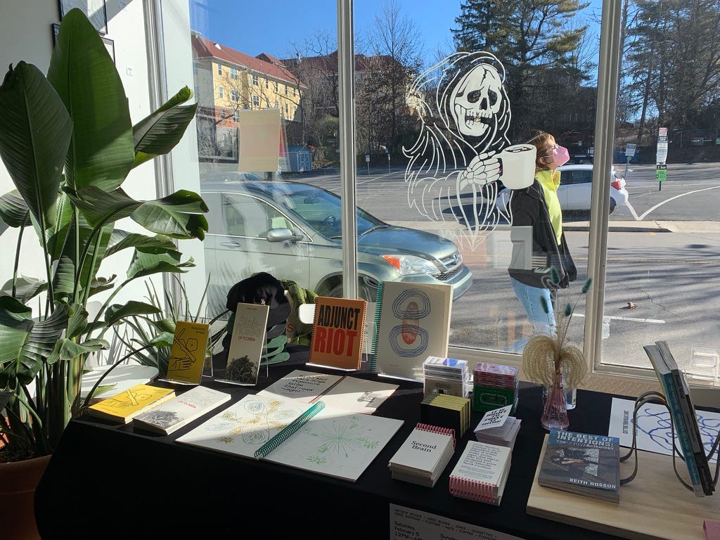 A vendor table full of artists' books in front of a sunny coffee shop window.