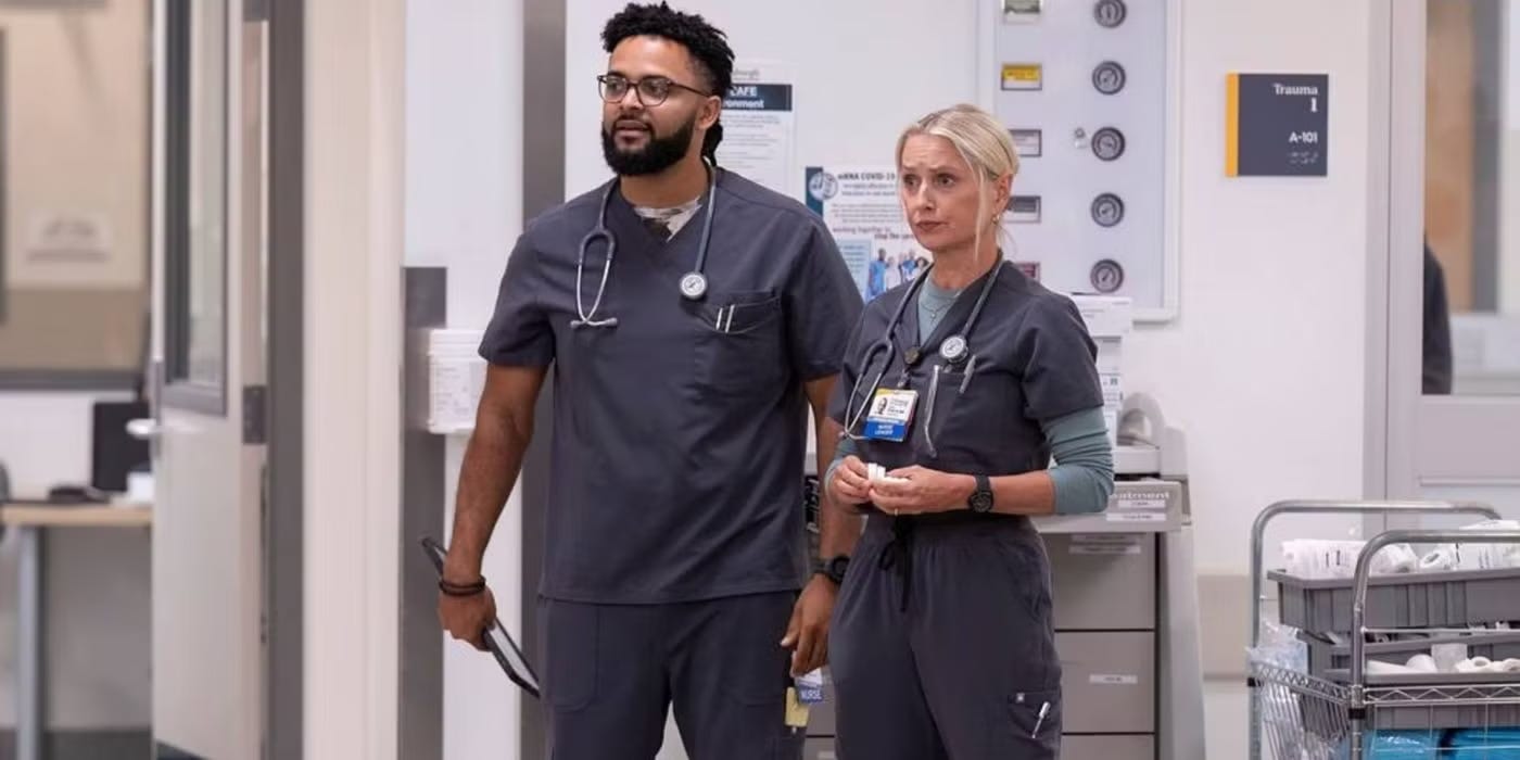 A photograph of two supporting characters from the television show The Pitt. At left, nurse Donnie is a large mixed-race black man with short braids, a beard, and glasses. At right, nurse Dana is a small white woman with blond hair pulled back in a bun. Both wear navy blue scrubs, stethoscopes around their necks, and serious facial expressions. They stand in what looks like an ER-setting, with carts of medical supplies in the background and a room labeled "Trauma 1" to the right of the frame.