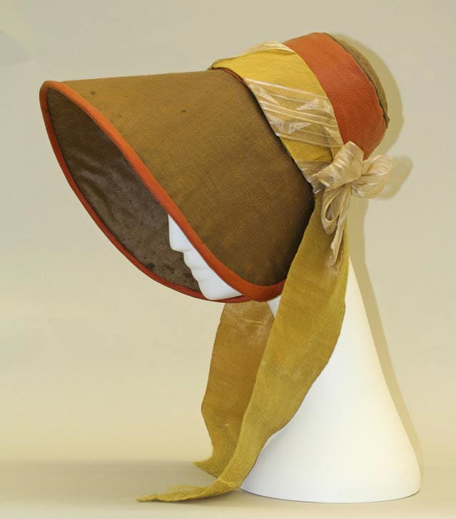 1830s bonnet displayed on a white mannequin head. The brown bonnet has burnt orange trim and ribbon on the back as well as a yellow ribbon just above the burnt orange at the back. This bonnet has sides that fully cover the sides of the face that look like blinders.