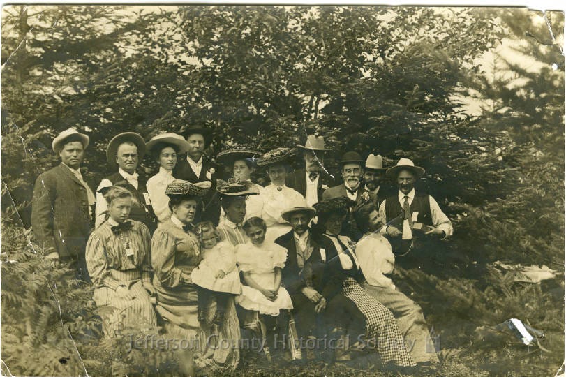 Historic outdoor group photograph of men, women, and children gathered among trees. Charles E. Coon stands in the back row, fourth from the right. Charles A. Pragge sits near the center of the front row, with young Helen “Nellie” Pragge next to him. Most other figures and the location are unidentified.