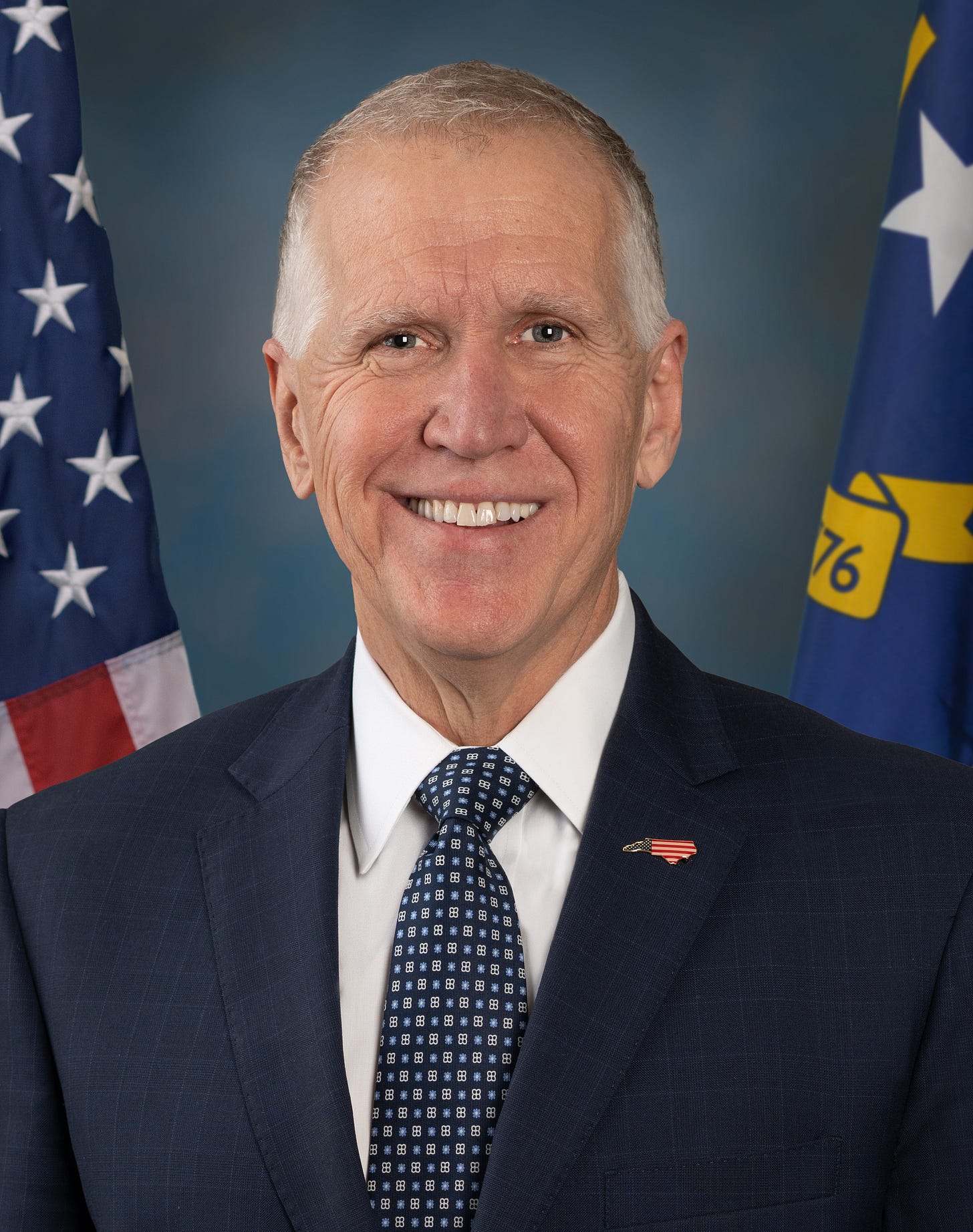 official portrait of Thom Tillis, smiling white haired balding white guy wearing a suit; flags of US and NC are behind him official portrait of Thom Tillis, smiling white haired balding white guy wearing a suit; flags of US and NC are behind him