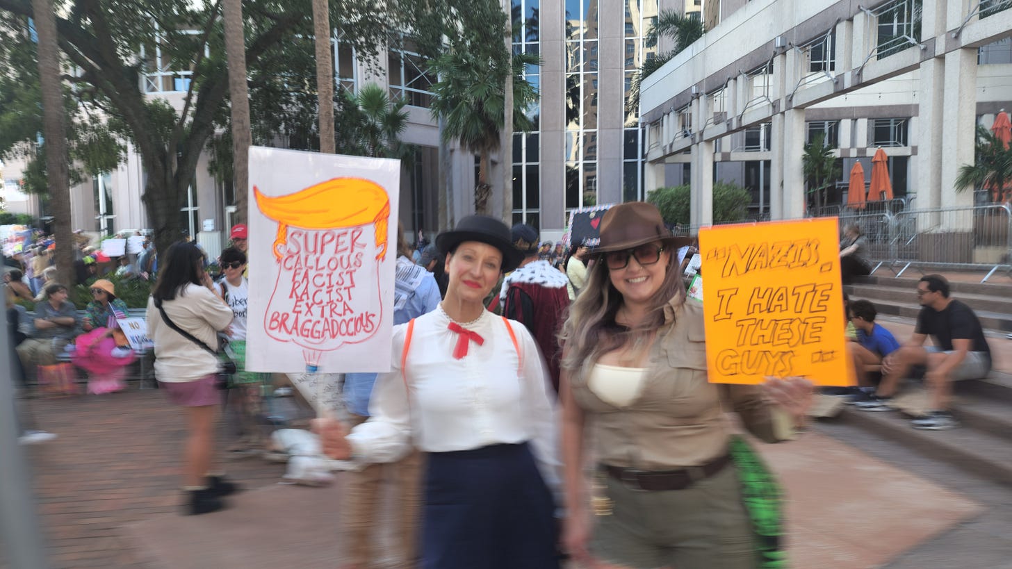 A slightly blurred, eye-level medium shot of two women in costume posing for the camera at an outdoor protest. The woman on the left is dressed as Mary Poppins, holding a sign that caricatures Donald Trump with the text "SUPER CALLOUS FACIST RACIST EXTRA BRAGGADOCIOUS." The woman on the right is dressed as Indiana Jones, smiling and holding an orange sign that reads, "'NAZIS,' I HATE THESE GUYS." Both are standing on a brick walkway, with other protestors and modern city buildings visible in the background.