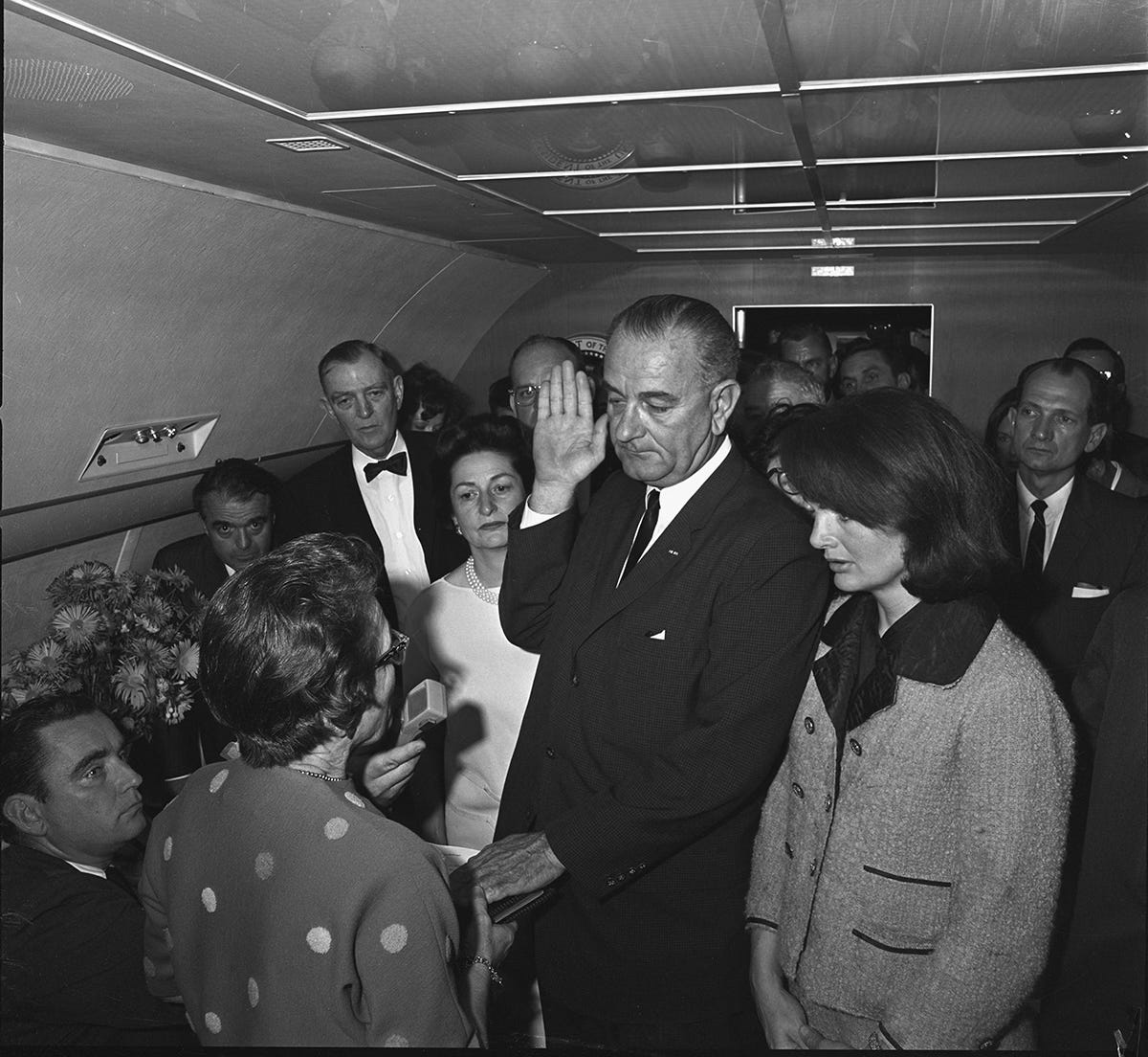 L-R: Mac Kilduff, Judge Sarah T. Hughes, Jack Valenti, Albert Thomas, Marie Fehmer (behind Thomas), Lady Bird Johnson, Chief Curry, President Lyndon B. Johnson, Evelyn Lincoln (eyeglasses only visible above LBJ's shoulder), Homer Thornberry (in shadow, partially obscured by LBJ), Roy Kellerman (partially obscured by Thornberry), Lem Johns (partially obscured by Mrs. Kennedy), Jacqueline Kennedy, Pamela Tunure (behind Brooks), Jack Brooks, Bill Moyers (mostly obscured by Brooks)