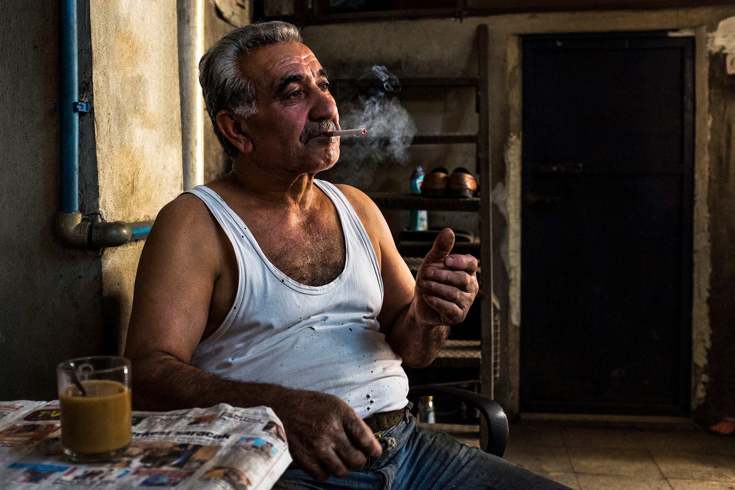 An older man in a white tank top sits indoors, smoking a cigarette. A glass of tea and a newspaper are on the table beside him. Warm light highlights his face, and the background is dimly lit and rustic.