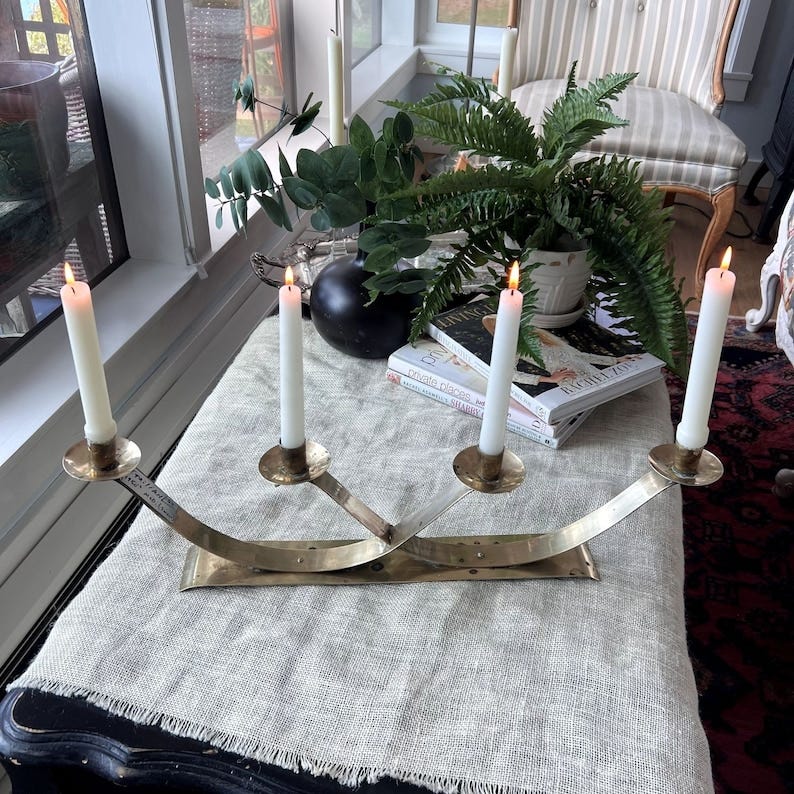 May include: Brass five-candle candelabra with four lit white taper candles. The candelabra has a curved, modern design and sits on a beige linen tablecloth.  Books with titles such as 'Create Places' and 'Rachel Zoe' are visible in the background, along with a potted fern plant.