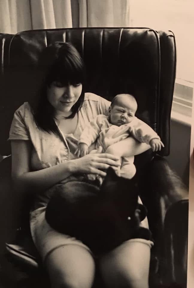 A black and white picture of a 24 year old Nina. She's sitting on a big leather chair with a nighty on and a newborn baby and black cat on her lap.